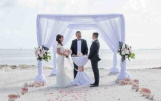 A bride and groom standing under a white wedding arch on the beach, beautifully arranged by key west wedding planners.