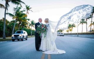 A bride and groom are seen standing on a road in Key West, surrounded by palm trees in the background.