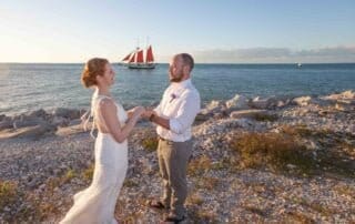 A bride and groom standing on the beach next to a sailboat, capturing stunning moments of their special day with the assistance of a key west wedding photographer.