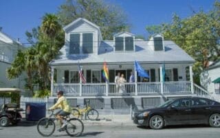A man on a bicycle rides past a white house with flags, capturing the scenic allure of Key West.