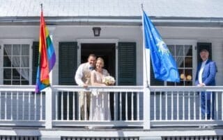 A bride and groom, accompanied by a key west wedding officiant, standing on the porch of a house decorated with rainbow flags for their beautiful wedding ceremony as organized by key west wedding planners.