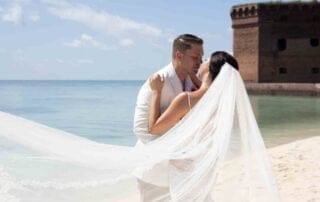 A bride and groom passionately kiss on the beach in front of an old fort, captured beautifully by a skilled Key West wedding photographer.