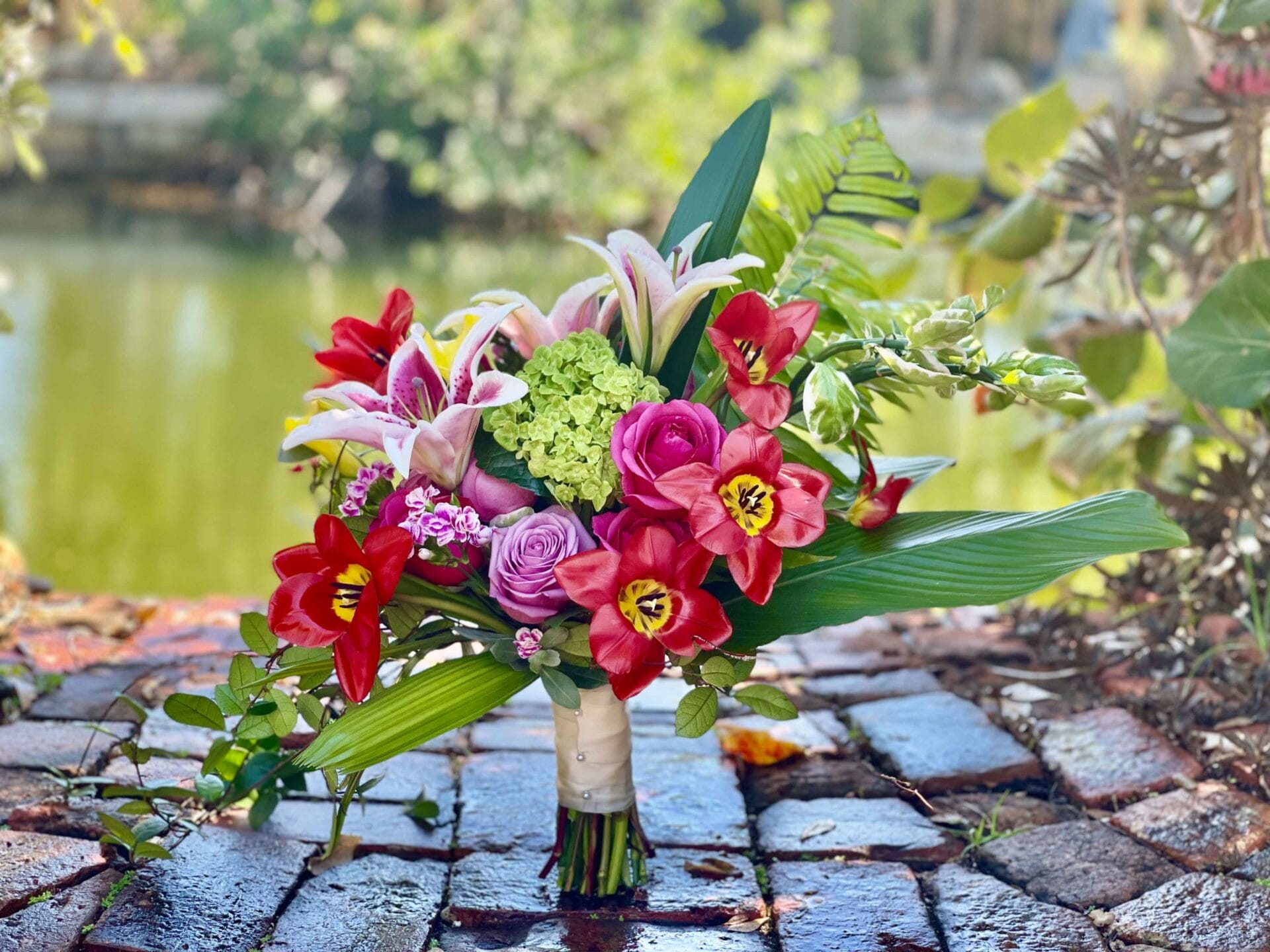 A bouquet of flowers sits on a brick walkway next to a pond at a beach wedding.
