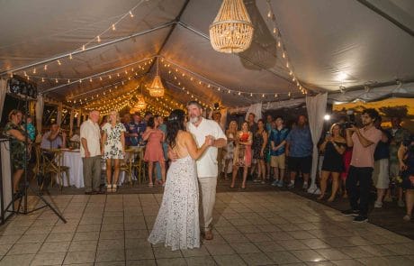 2023-11-17 Andrea and Pete Wedding, Ocean Key Resort and Spa, Key West Part 3 Social Media -3903 This image captures a joyous wedding reception at the Ocean Key Resort and Spa in Key West. The bride and groom are in the center of the dance floor, sharing a tender moment, surrounded by guests. The venue is adorned with string lights creating a warm, inviting glow that complements the dusky evening sky visible through the open sides of the tent. The scene is lively yet intimate, with the elegant attire of the guests adding to the festive atmosphere. The oceanfront setting, combined with the sophisticated decor, offers a glimpse into a memorable celebration at this destination wedding.
