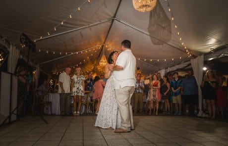 2023-11-17 Andrea and Pete Wedding, Ocean Key Resort and Spa, Key West Part 3 Social Media -3905 This image captures a joyous wedding reception at the Ocean Key Resort and Spa in Key West. The bride and groom are in the center of the dance floor, sharing a tender moment, surrounded by guests. The venue is adorned with string lights creating a warm, inviting glow that complements the dusky evening sky visible through the open sides of the tent. The scene is lively yet intimate, with the elegant attire of the guests adding to the festive atmosphere. The oceanfront setting, combined with the sophisticated decor, offers a glimpse into a memorable celebration at this destination wedding.