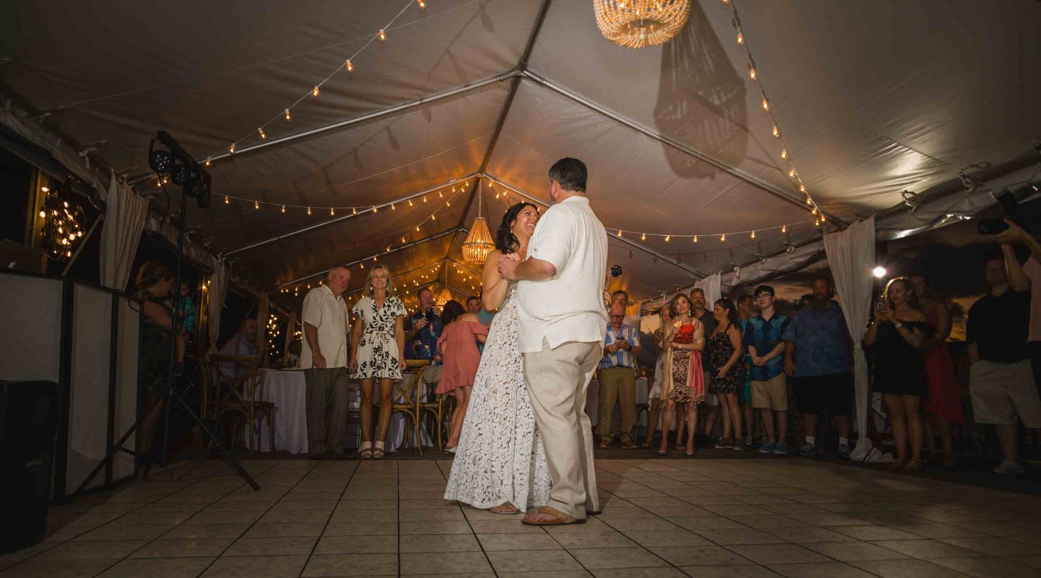 This image captures a joyous wedding reception at the Ocean Key Resort and Spa in Key West. The bride and groom are in the center of the dance floor, sharing a tender moment, surrounded by guests. The venue is adorned with string lights creating a warm, inviting glow that complements the dusky evening sky visible through the open sides of the tent. The scene is lively yet intimate, with the elegant attire of the guests adding to the festive atmosphere. The oceanfront setting, combined with the sophisticated decor, offers a glimpse into a memorable celebration at this destination wedding.