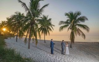 A serene sunrise wedding at Smathers Beach, Key West, featuring a couple exchanging vows on the sandy shore. Palm trees frame the golden sunrise, creating a picturesque and romantic setting. The calm ocean and the soft morning light provide a perfect backdrop for intimate nuptials.