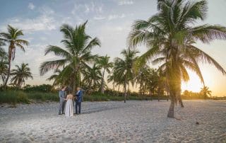 A serene sunrise wedding at Smathers Beach, Key West, featuring a couple exchanging vows on the sandy shore. Palm trees frame the golden sunrise, creating a picturesque and romantic setting. The calm ocean and the soft morning light provide a perfect backdrop for intimate weddings.
