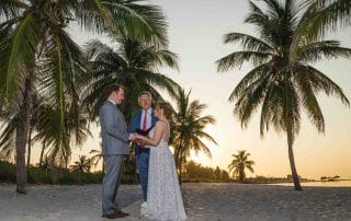 A bride and groom standing on the beach with palm trees in the background.