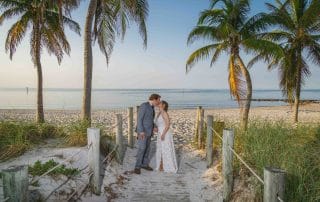 A serene sunrise wedding at Smathers Beach, Key West, featuring a couple exchanging vows on the sandy shore. Palm trees frame the golden sunrise, creating a picturesque and romantic setting. The calm ocean and the soft morning light provide a perfect backdrop for intimate weddings.
