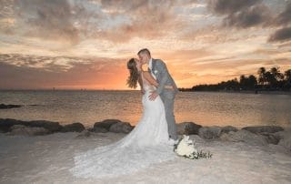 A couple in wedding attire kissing on a beach at sunset, with the ocean and palm trees in the background.
