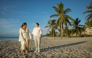 Two women walking hand in hand on a sandy beach, with palm trees and buildings in the background during early morning light, illustrating the beauty of inclusive wedding planning in Key West.