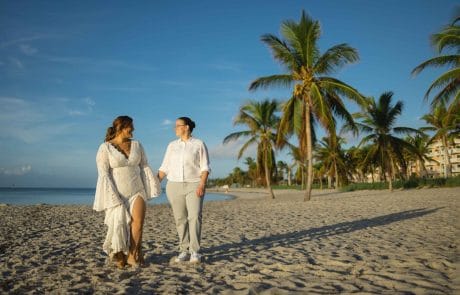 2022-11-26 Maria and Brittany Sunrise Smathers Beach Wedding Social media -3-144 Two women walking hand in hand on a sandy beach, with palm trees and buildings in the background during early morning light, illustrating the beauty of inclusive wedding planning in Key West.