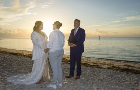 2022-11-26 Maria and Brittany Sunrise Smathers Beach Wedding Social media -3-57 A couple exchanges vows at a beachside wedding ceremony during sunset in Key West, officiated by a man holding a book.