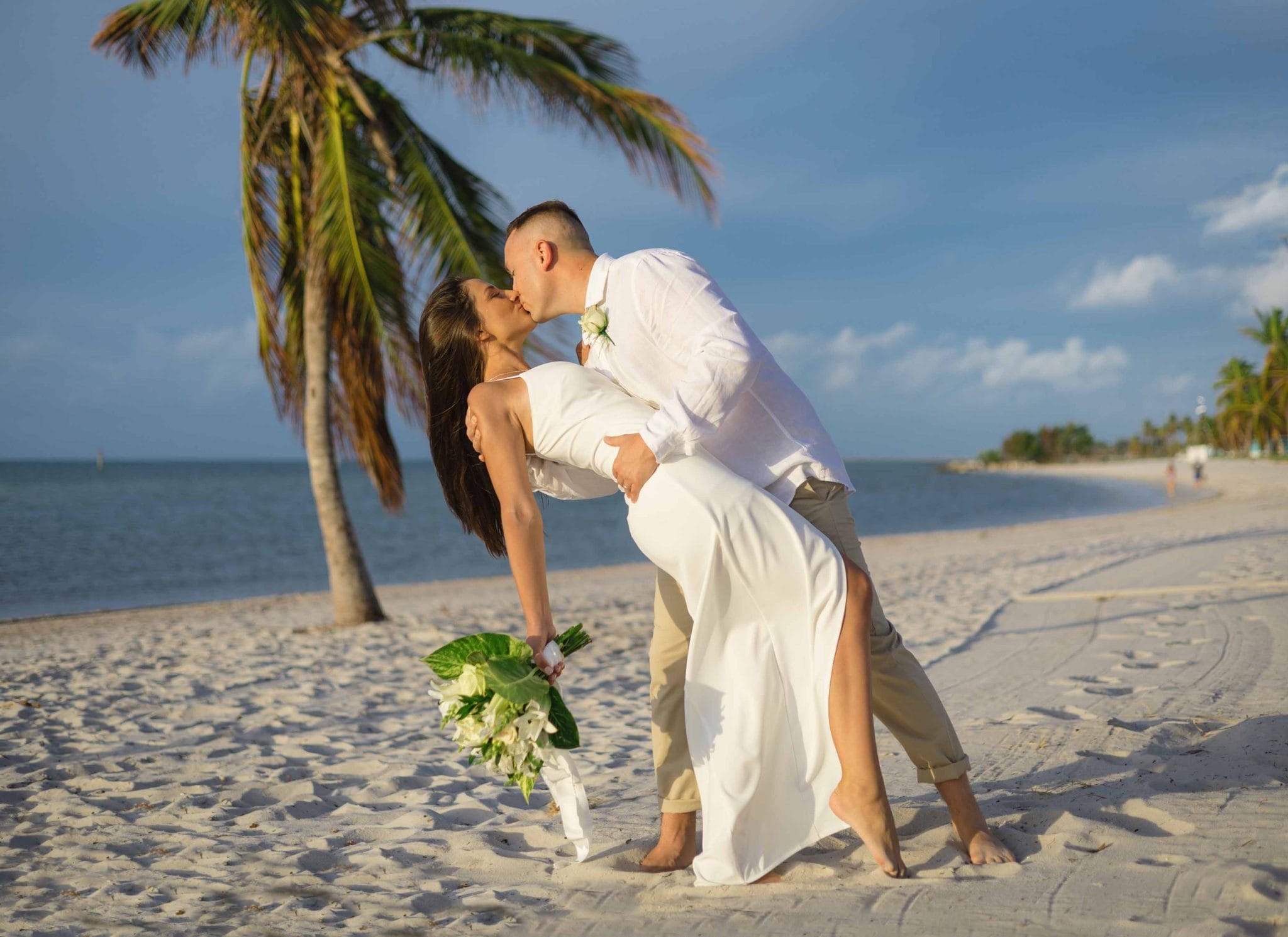 A couple in wedding attire kissing on a sandy beach under a palm tree, with clear blue skies and ocean in the background.
