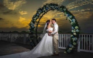 A bride and groom kissing under a floral arch, with a dramatic Key West sunset sky in the background.