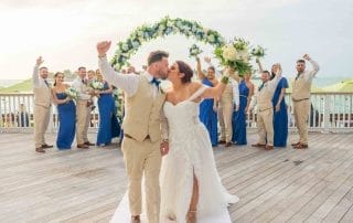 A bride and groom kiss on a wooden pier with their wedding party, wearing blue and beige, cheering behind them under a floral arch.