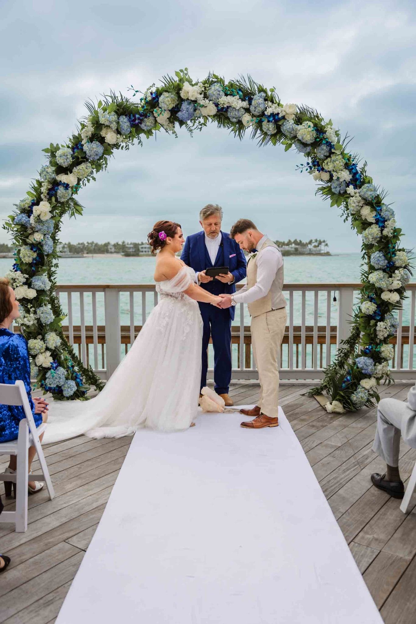 2024-01-13 Proofing Album Part 1-5201 A bride and groom exchange rings at an outdoor wedding ceremony by the sea, under a floral archway, with an officiant and guests seated nearby.
