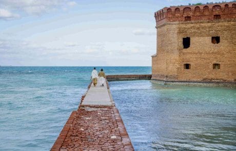 2024-01-29 Dry Tortugas Wedding Social media-8286 Two people walking towards a large brick fort by the sea, connected by a narrow brick walkway under a cloudy sky.