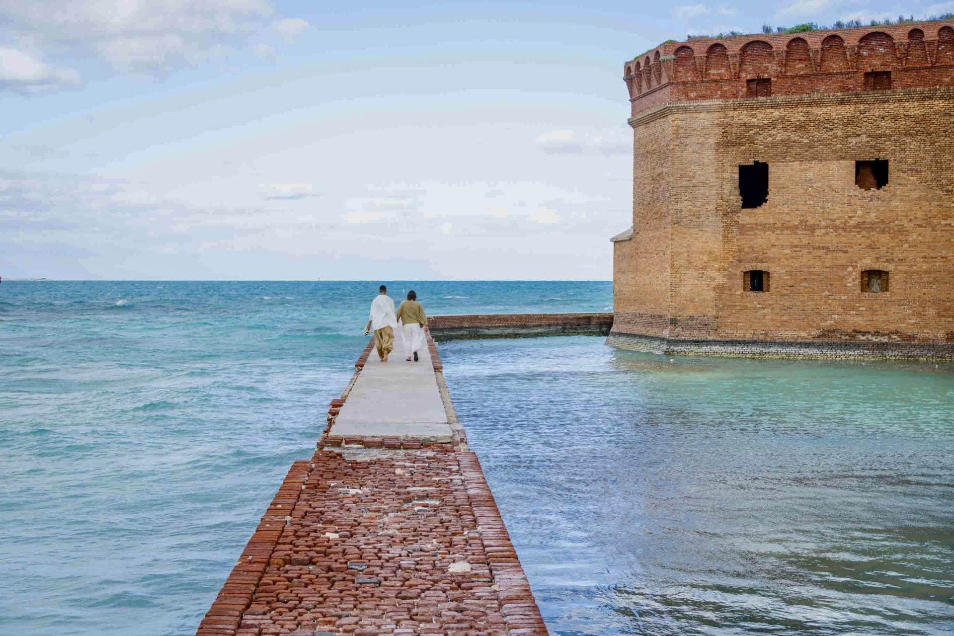 2024-01-29 Dry Tortugas Wedding Social media-8286 Two people walking towards a large brick fort by the sea, connected by a narrow brick walkway under a cloudy sky.