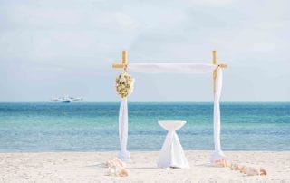A simple wedding arch decorated with white drapes and floral arrangements stands on a sandy beach overlooking a calm sea.