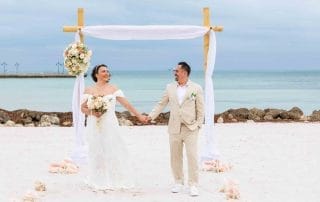 A happy couple holding hands under a simple wedding arch on a sandy beach in Key West, with a cloudy sky and calm sea in the background.