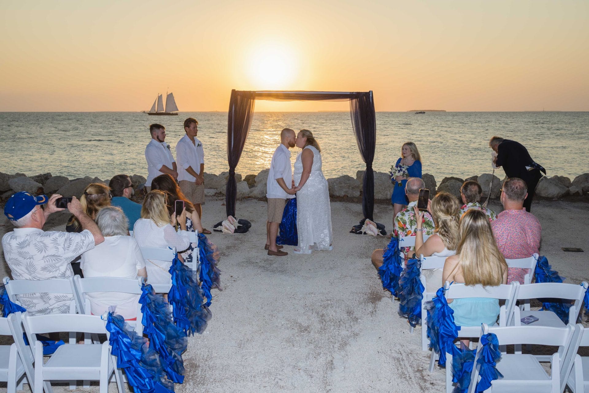 A couple kisses under a canopy at a beach wedding ceremony, with guests seated around and a sailboat visible during sunset.