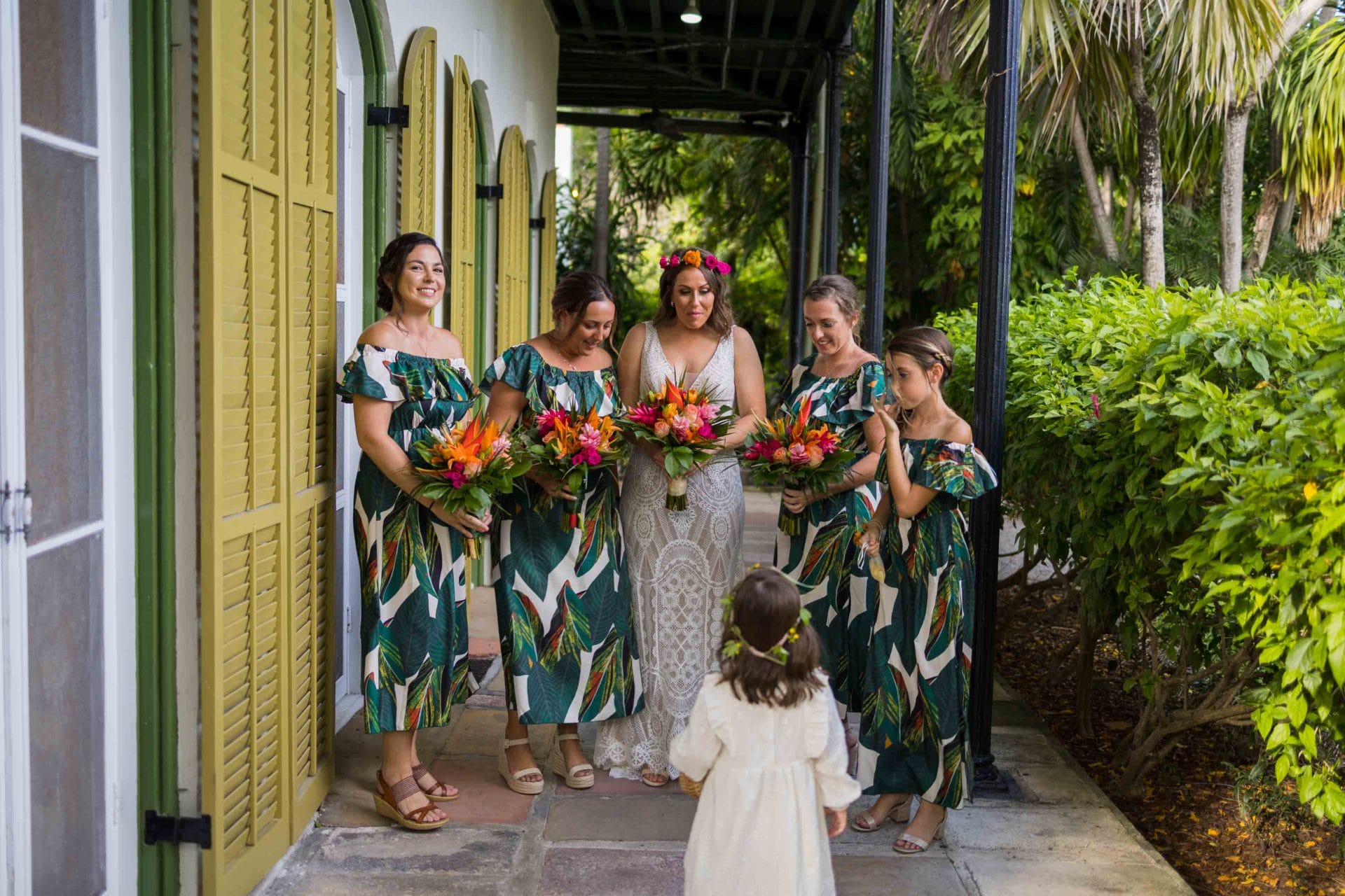 Wedding at Hemingway House, Key West Bride and bridesmaids in matching tropical dresses holding colorful bouquets walking down a garden path, greeted by a young flower girl.