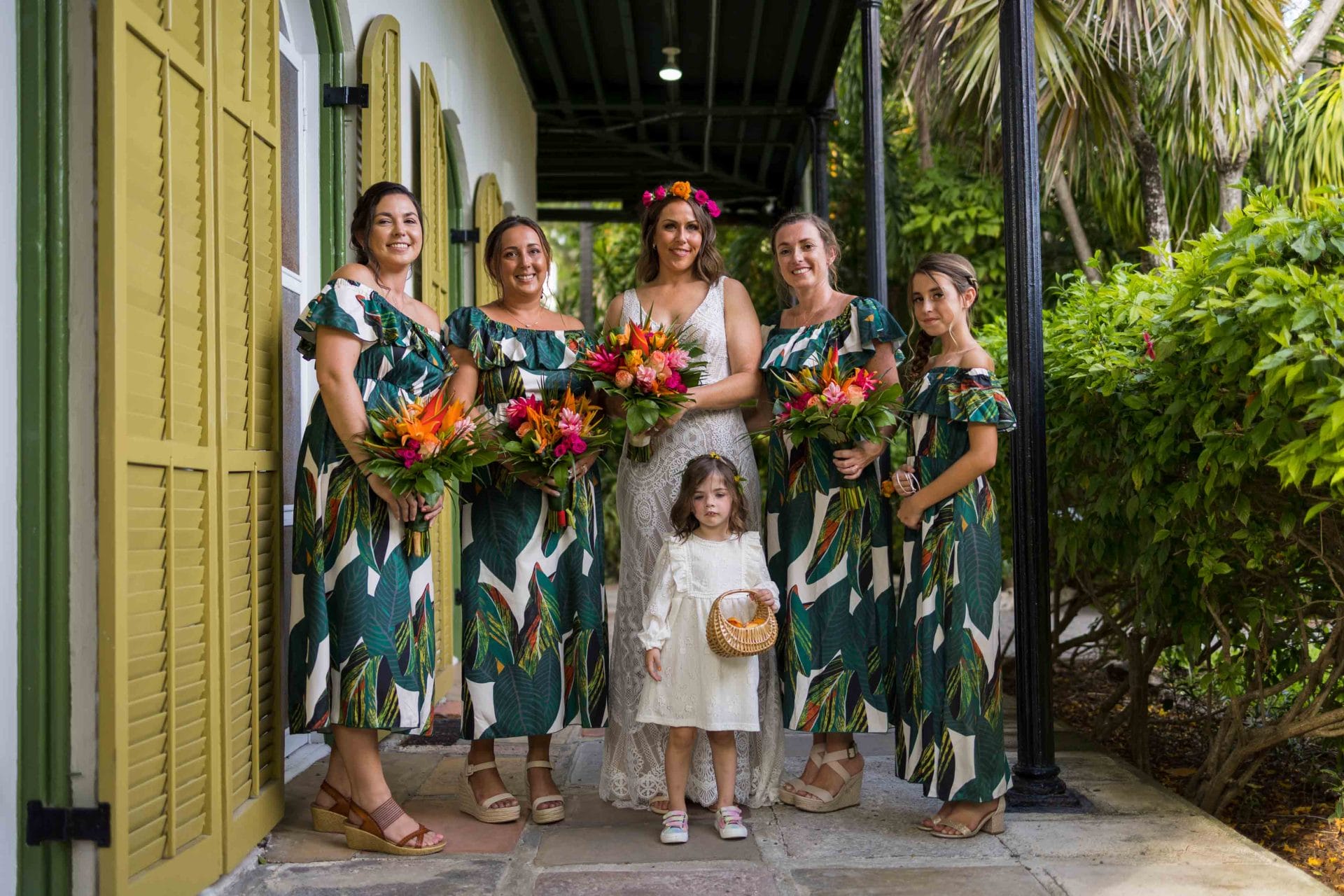 A bride and her bridesmaids, wearing matching tropical dresses, pose together with a young flower girl in a garden pathway.