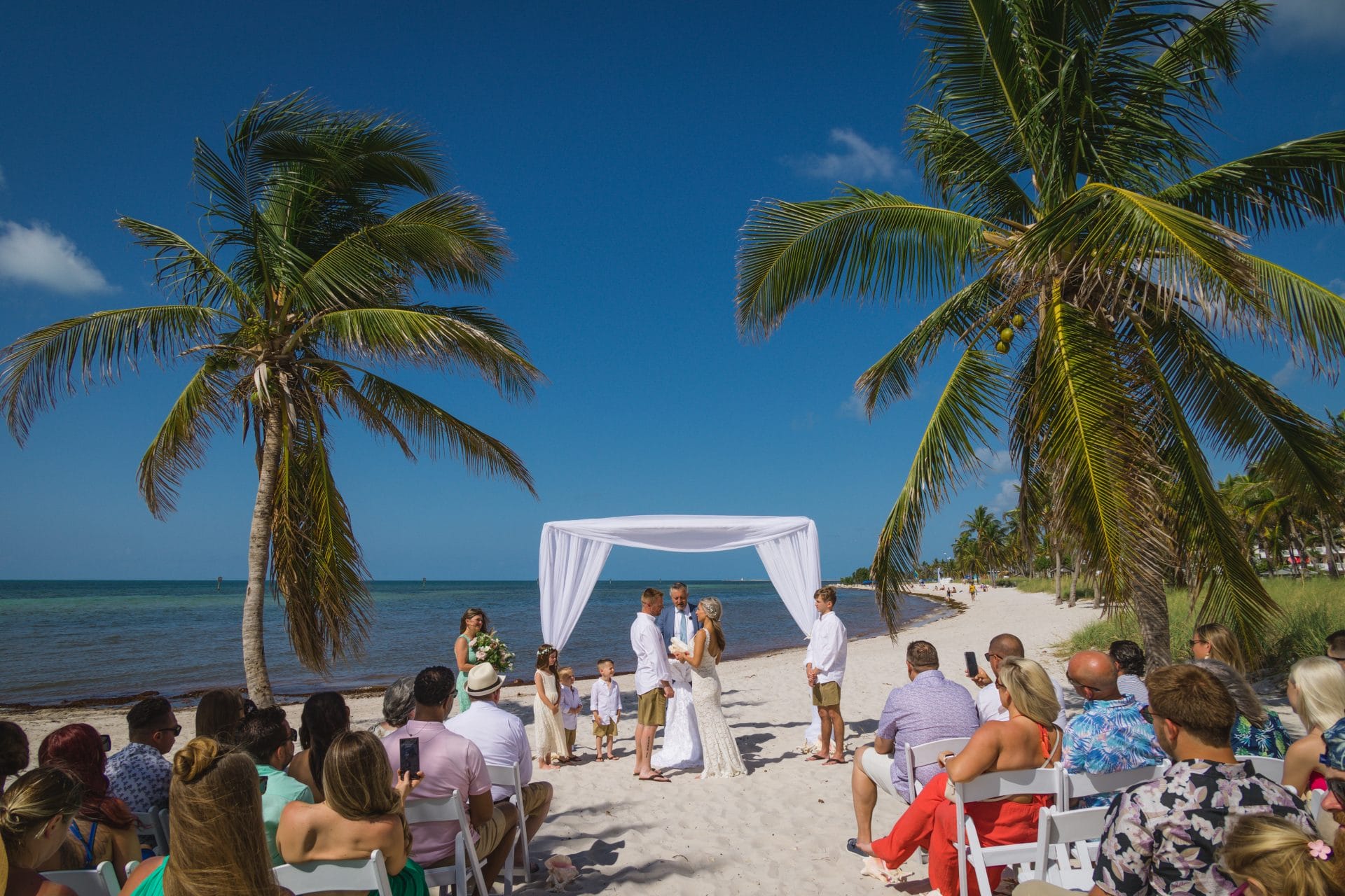 2023-04-21 Amber and Douglas Smathers Beach -1393 A beach wedding ceremony with a couple under a white canopy, guests seated on either side, palm trees nearby, and a clear blue sky overhead.