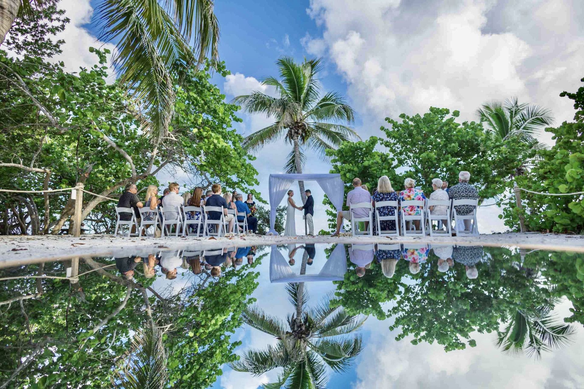 A beach wedding ceremony reflected in a pool, showing a couple exchanging vows under an arch, surrounded by guests and palm trees.