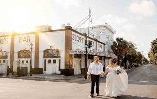 A newlywed couple holding hands and walking across a sunny street, with "sloppy joe's bar" in the background.