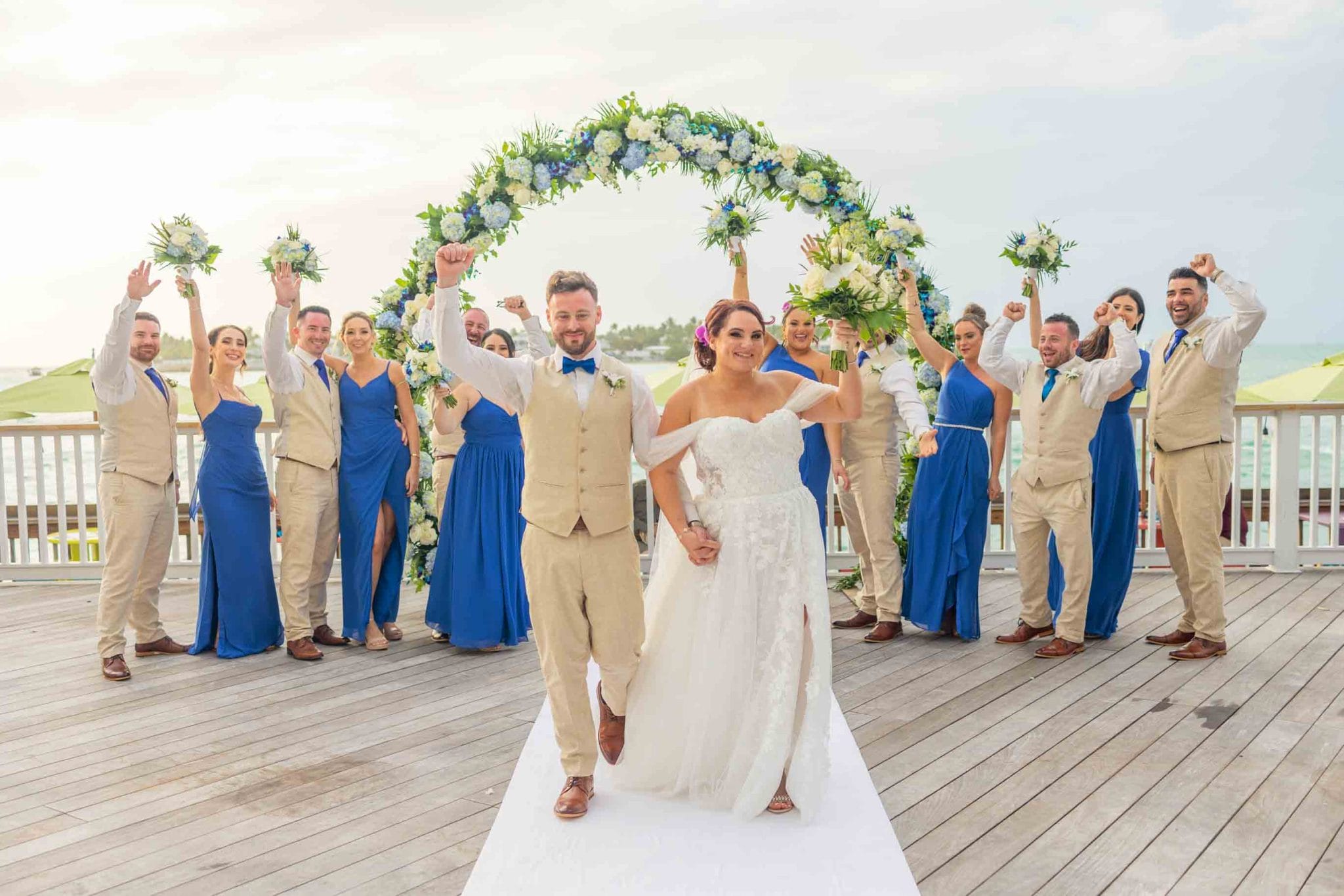 Bride and groom walking down the aisle under a floral arch, flanked by bridesmaids and groomsmen in blue, celebrating on a sunny waterfront platform.