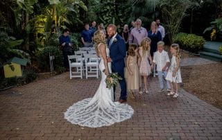 Bride and groom hold hands in a garden setting, surrounded by guests and four young children during a wedding ceremony.