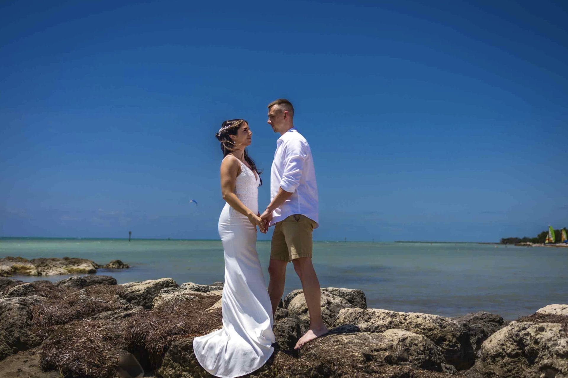 2024-04-21 Farrah and Kevin Proofing Album-2-116 A couple, dressed in white, standing on rocks by the sea under a clear blue sky, holding hands and looking at each other. Capture your love story with the Key West Photo Adventure Package for an unforgettable memory.