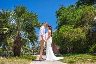 A couple stands face to face, holding hands outdoors on a sunny day. The man wears a white shirt and shorts, and the woman wears a white dress. Surrounded by lush greenery and palm trees, they embrace the moment as part of their *Capture Your Love Story: Key West Photo Adventure Package*.