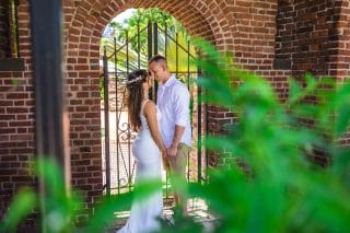 A couple stands in front of an archway with wrought iron gates, holding hands and facing each other. The woman wears a white dress, and the man wears a white shirt and beige shorts. Greenery is in the foreground—perfect for those looking to Capture Your Love Story as part of the Key West Photo Adventure Package.
