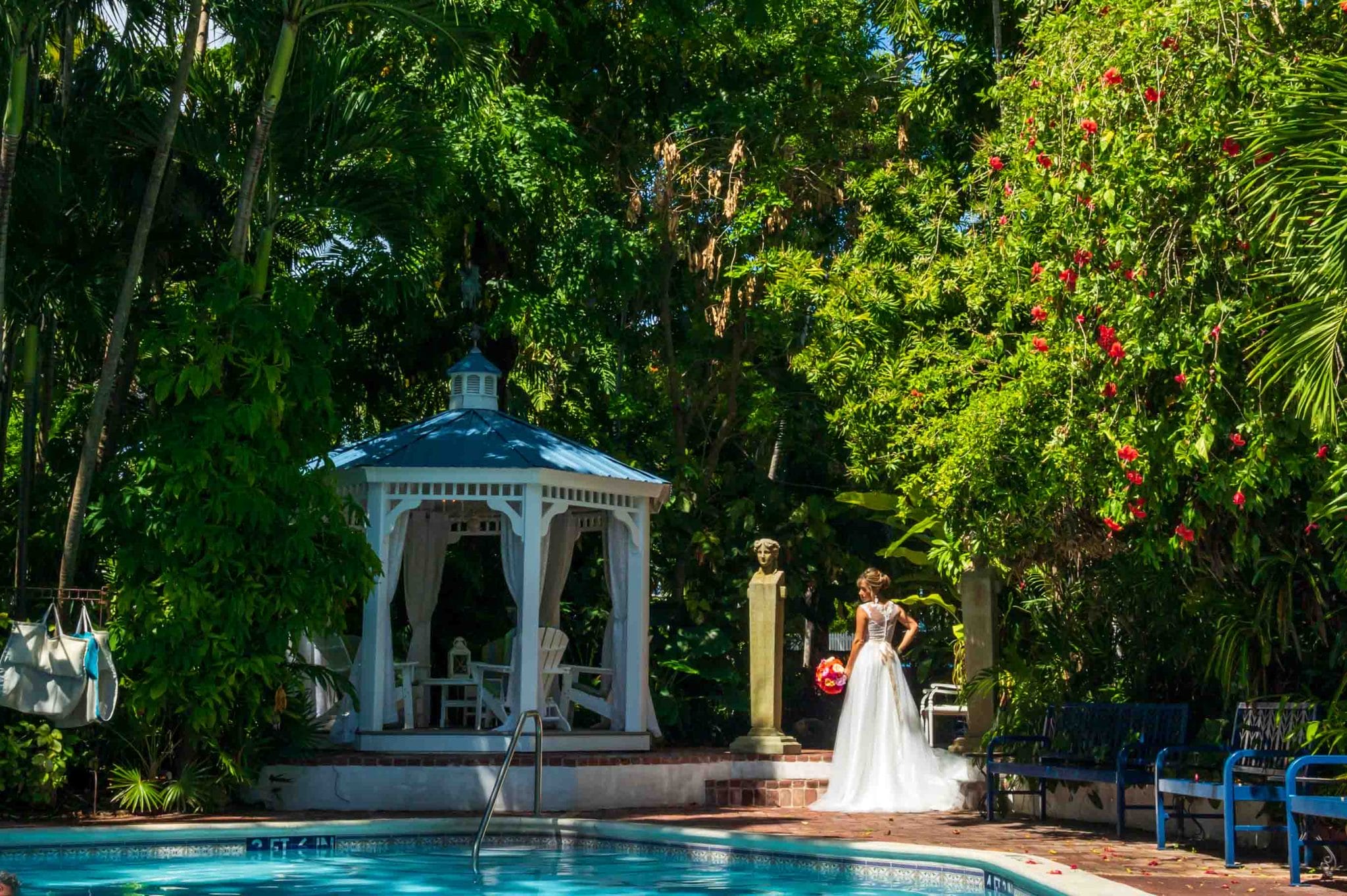 Garden’s Hotel Wedding A bride in a white dress stands near a gazebo by a pool, surrounded by lush greenery and red flowers, in a serene garden setting.