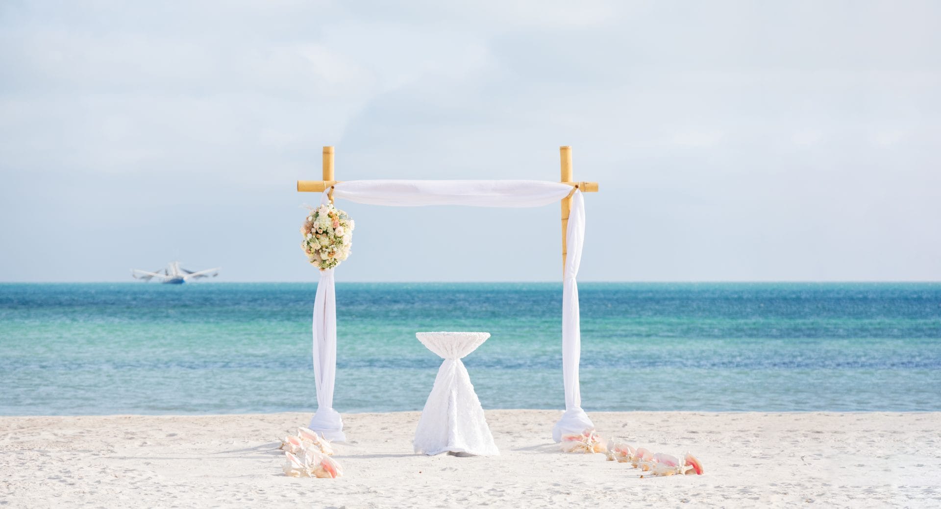 A simple wedding arch decorated with white fabric and flowers stands on a sandy beach, overlooking a calm sea with a distant boat.