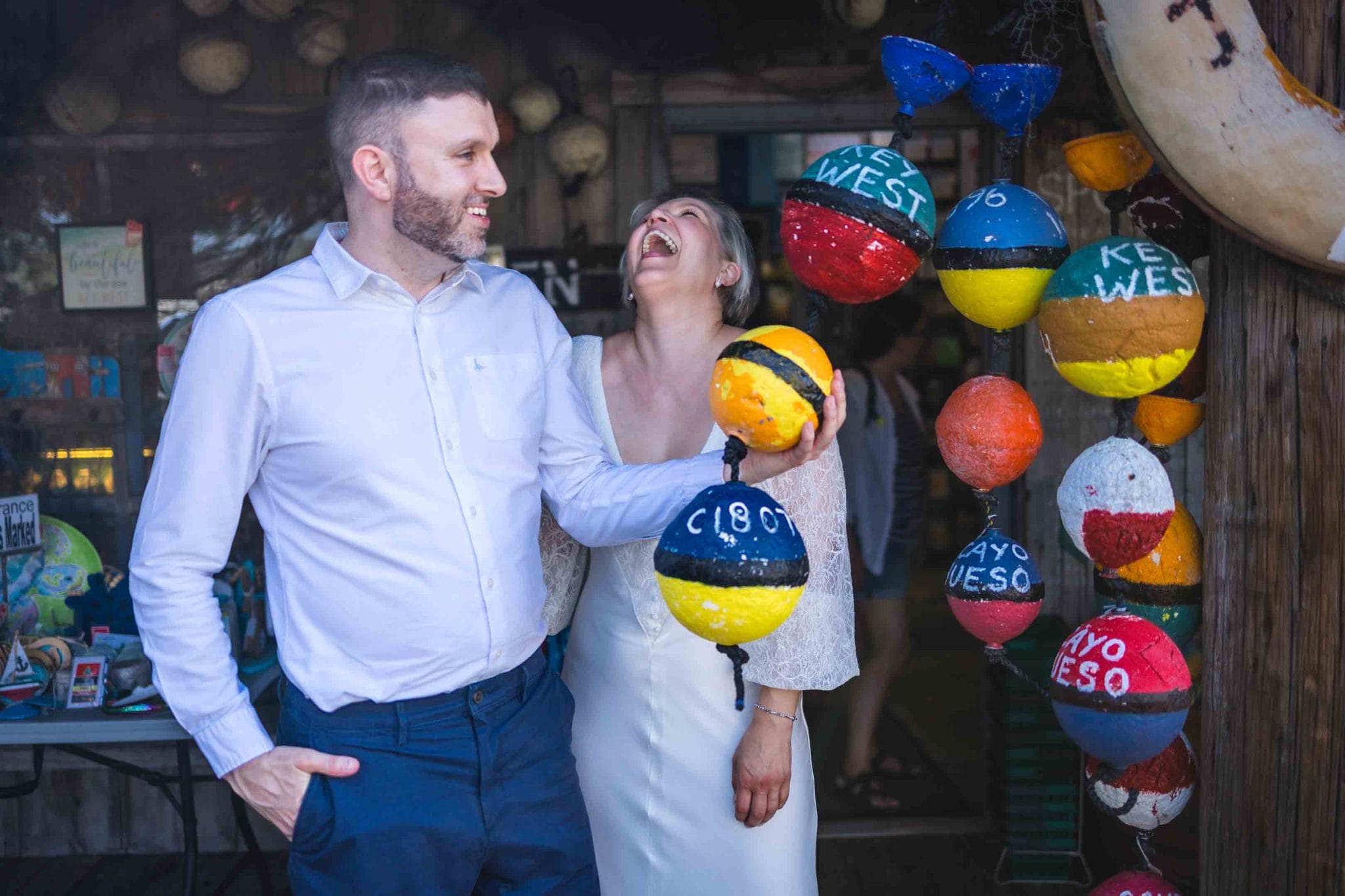Fort Zachary Taylor Park Wedding, Key West A smiling couple stands by colorful buoy decorations, with the words "Key West" visible, while the woman laughs joyfully.