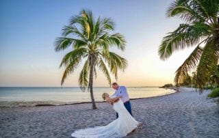 A couple in wedding attire poses under a palm tree on a sandy beach at sunset, with the ocean in the background.