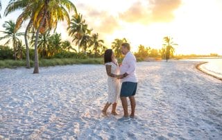 A couple stands on a sandy beach at sunset, surrounded by palm trees, holding hands and facing each other.