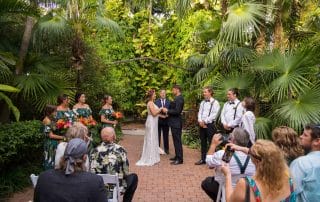 A couple stands at the altar in a tropical garden setting, surrounded by bridesmaids and groomsmen. Guests are seated, some taking photos of the ceremony.