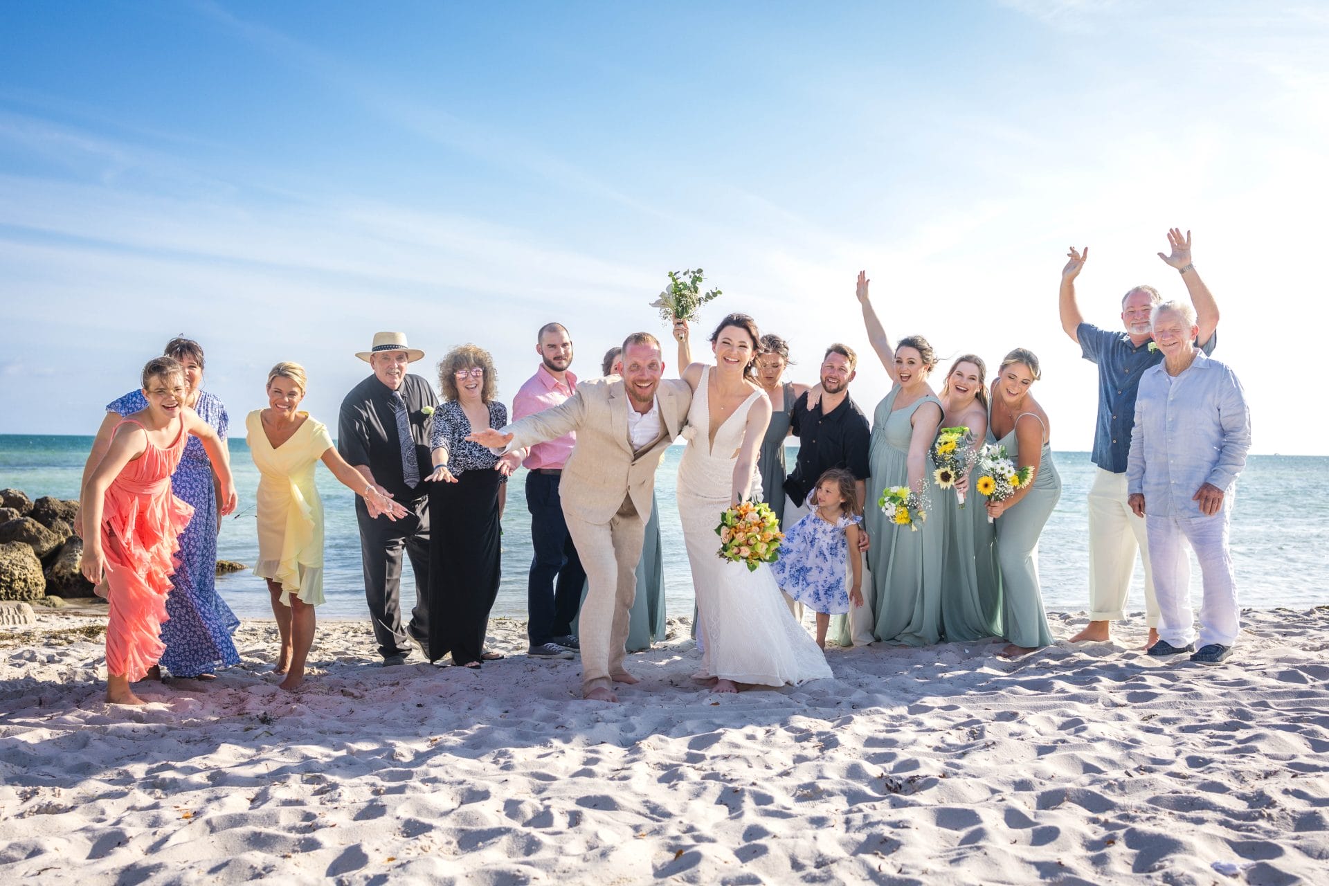 2024-03-14SmathersBeachWeddingKeyWest-4771 copy A wedding party poses joyfully on a sandy beach with a blue sky and ocean in the background. The bride and groom stand in the center, surrounded by friends and family.