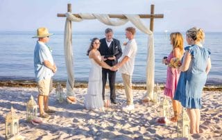 A couple exchanges vows at a beach wedding ceremony, surrounded by a small group of attendees. The backdrop features the ocean, a wooden arch, and lanterns on the sand.