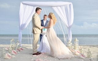 A couple stands under a decorated canopy on a sandy beach, holding hands during a wedding ceremony, with an officiant and the ocean in the background.
