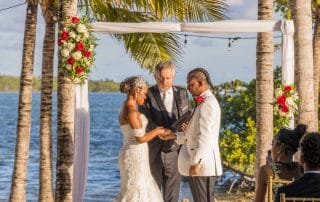 A couple stands under a floral arch during a beach wedding ceremony, with a man officiating in the center. Guests are seated on the right. Palm trees and water are in the background.