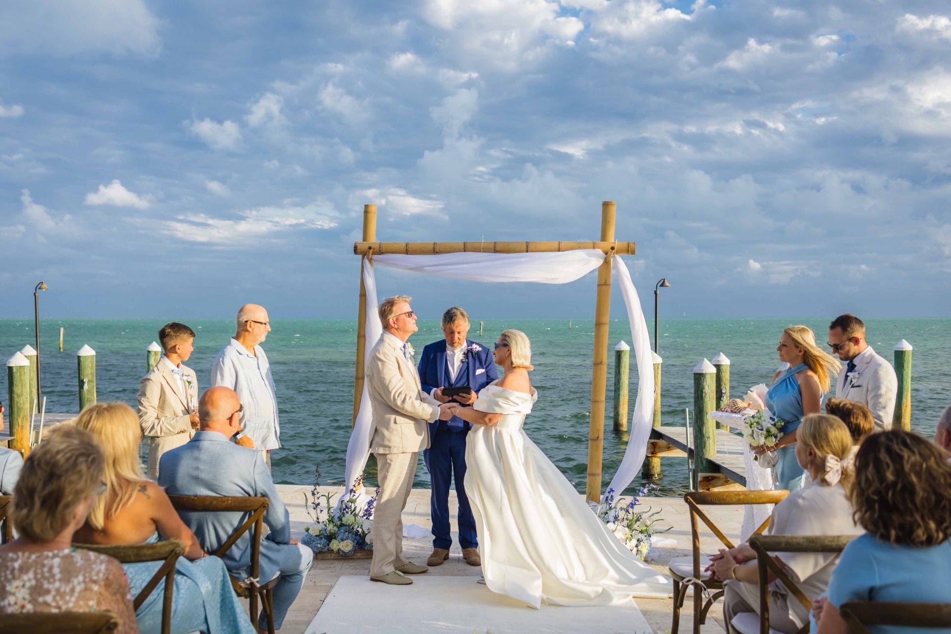 2024-10-29 SOcial media_-0377 A couple exchanges vows under a bamboo arch by the ocean, surrounded by guests, with chairs set up on a dock. The sky is partly cloudy.