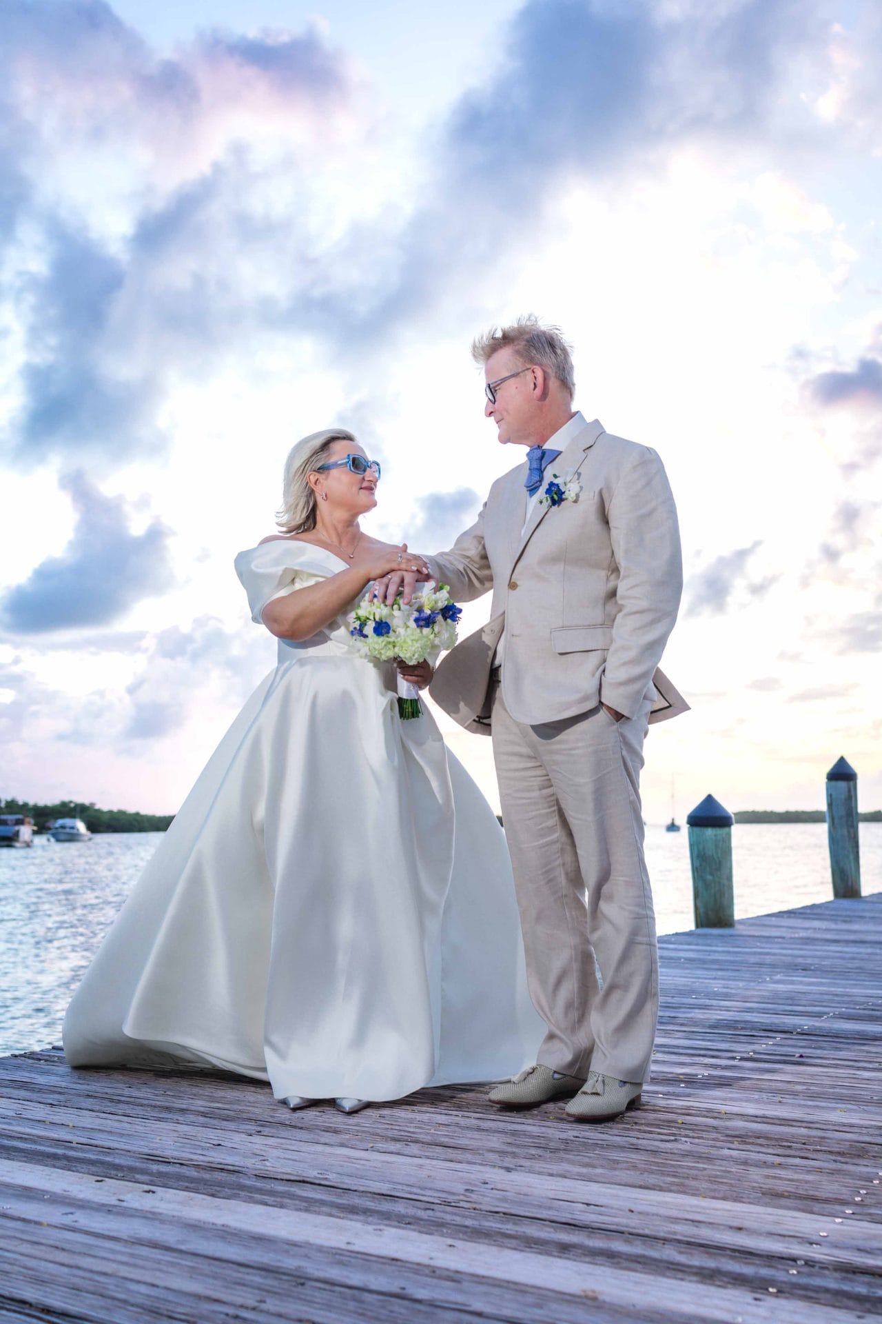 2024-10-29 SOcial media_-0687 A couple in wedding attire stands on a wooden dock by the water, with a cloudy sky in the background. The bride holds a bouquet, and they gaze at each other.