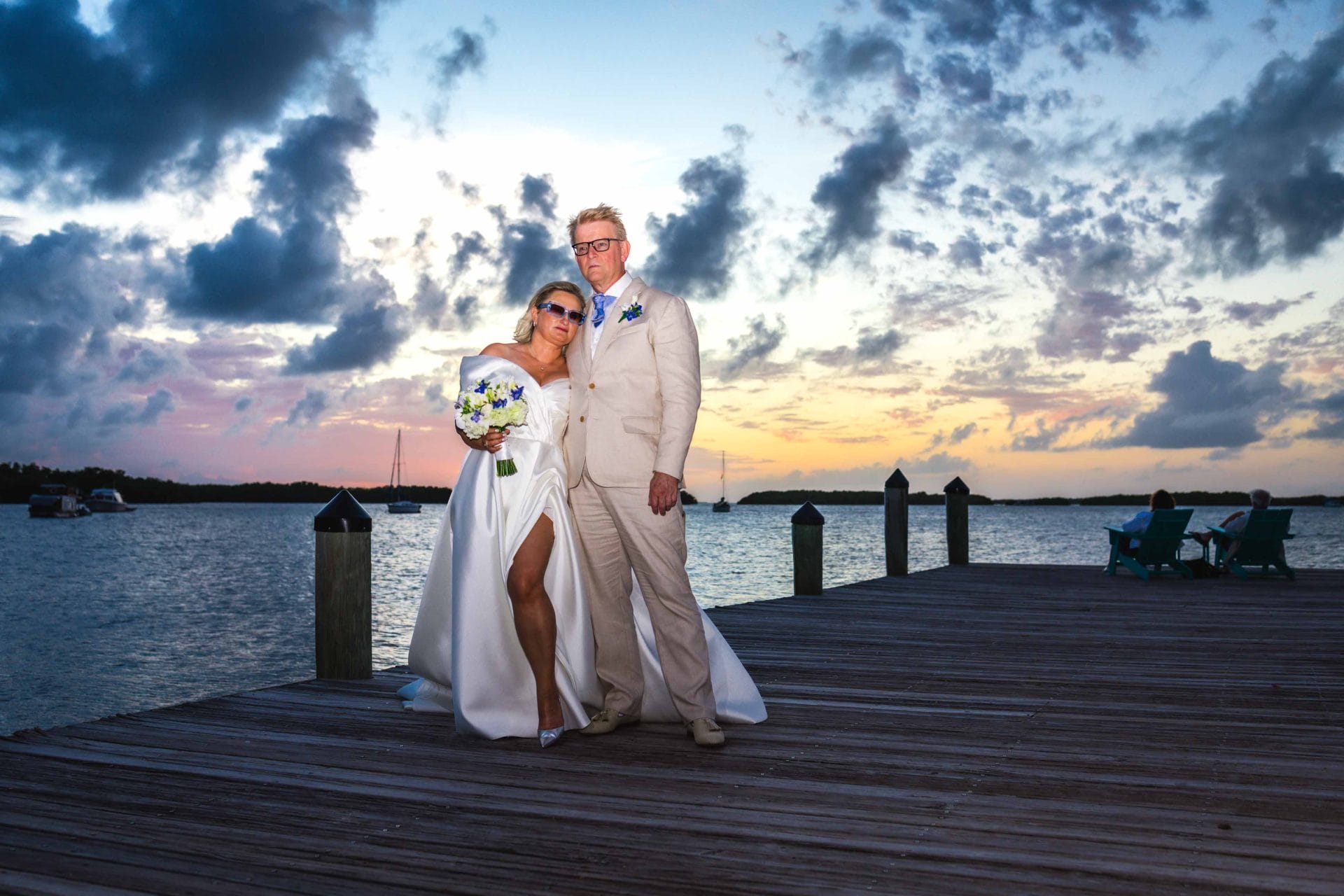 2024-10-29 SOcial media_-0717 Bride and groom pose on a wooden dock at sunset, with a scenic view of clouds, water, and distant boats in the background.
